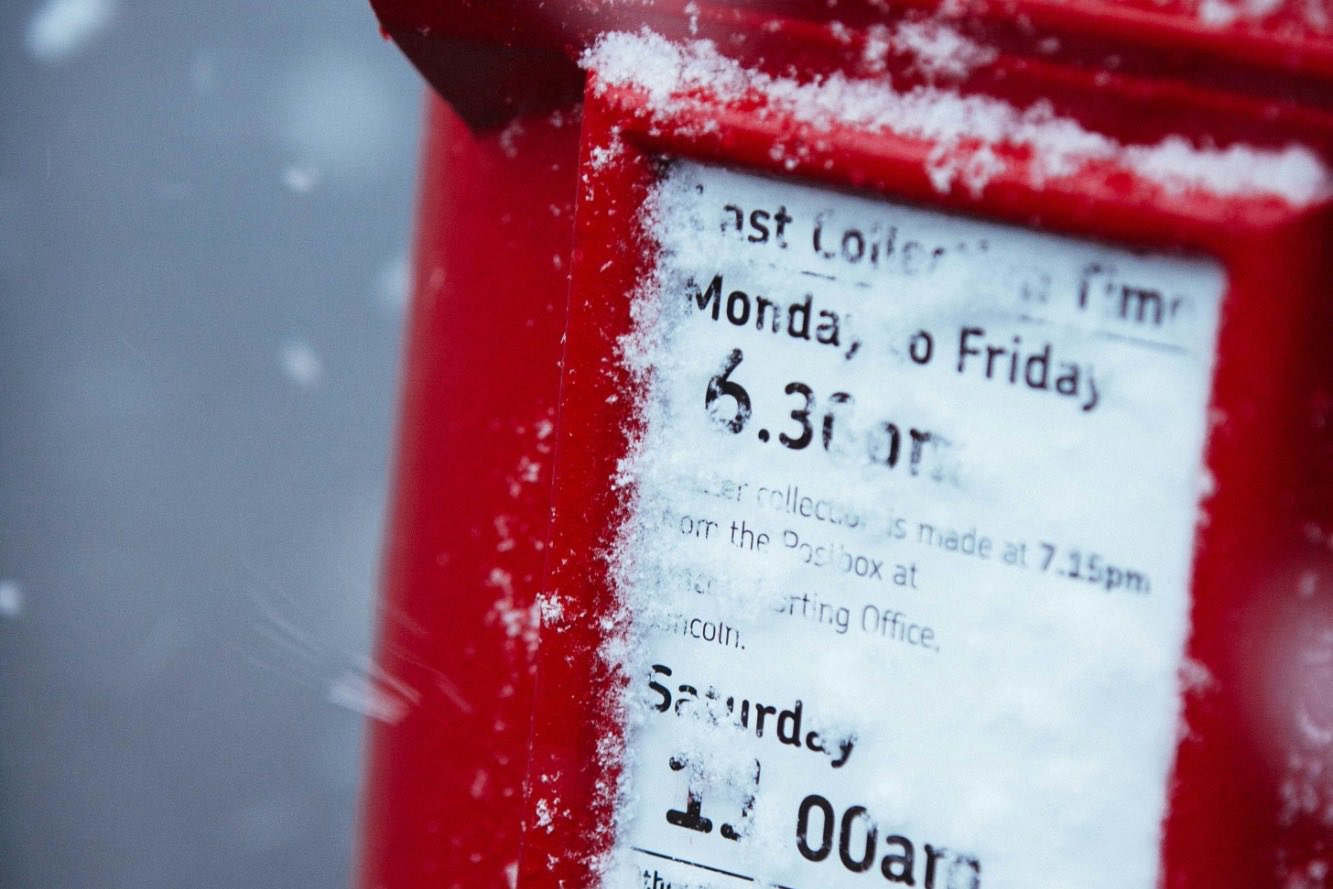 A red Royal Mail postbox dusted with snow, showing its last collection times